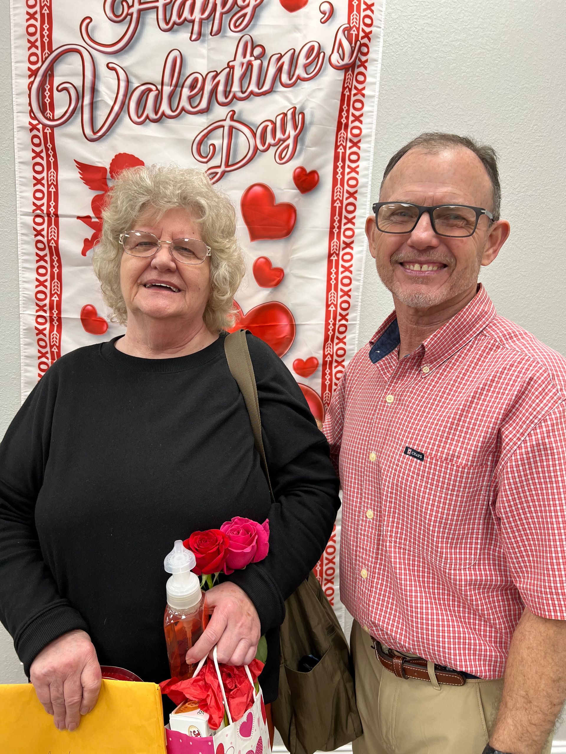 Woman with roses and gift bag, and man with glasses, standing in front of a Valentine's Day banner.