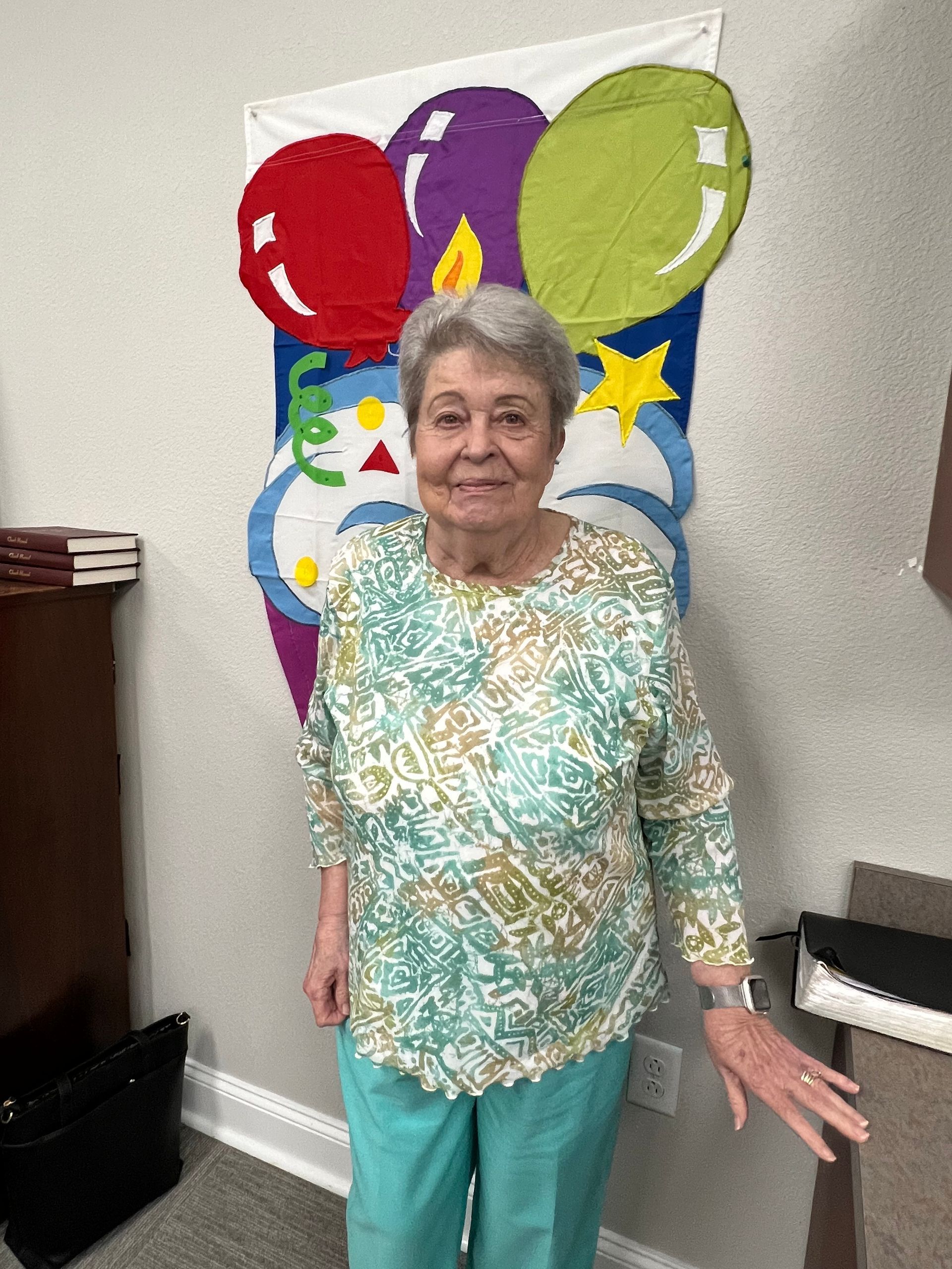 Woman in patterned top and teal pants smiles in front of a birthday-themed banner with balloons.