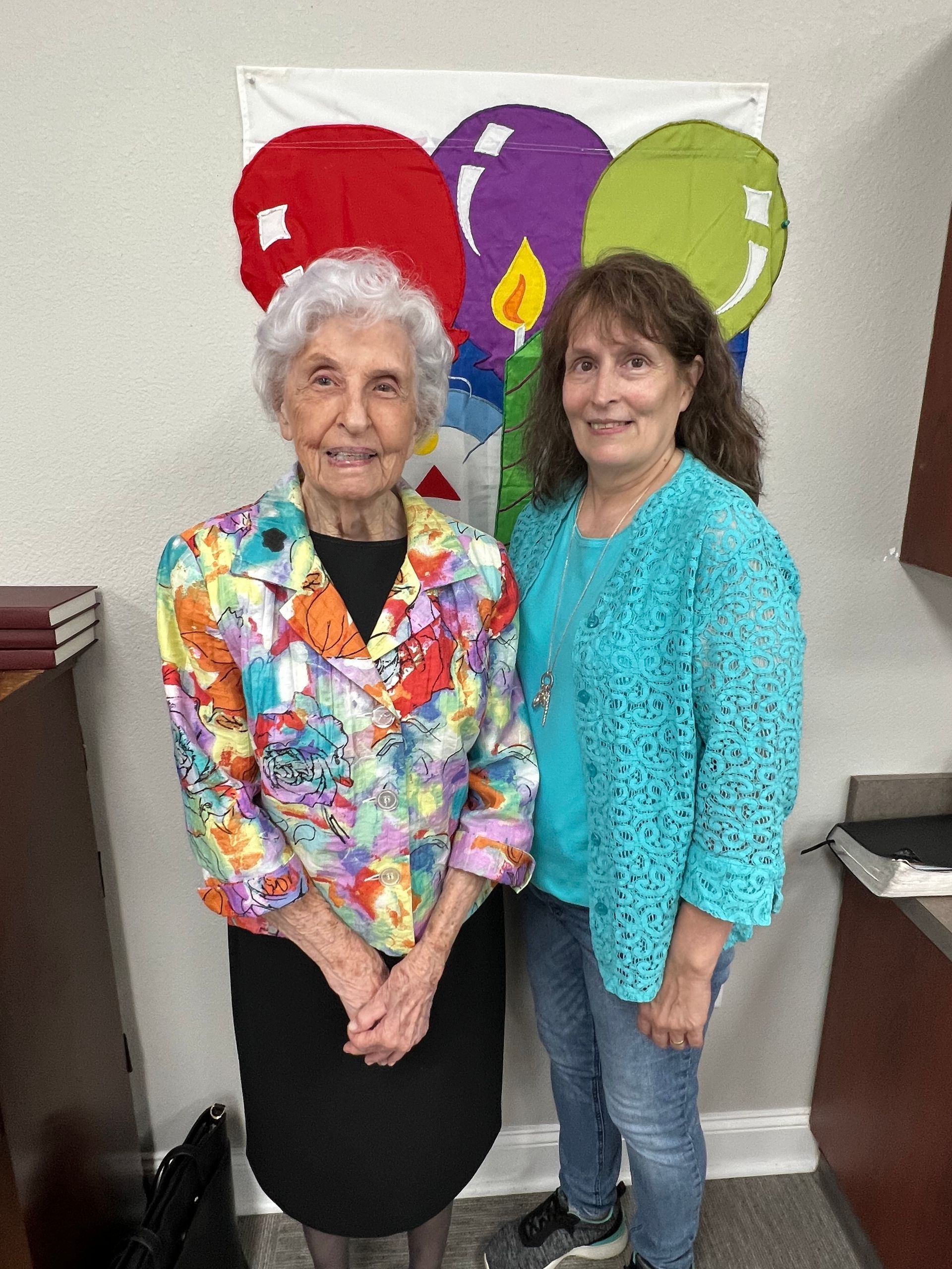 Two women stand smiling in front of a birthday banner with balloons. One wears a colorful jacket, the other a blue sweater.