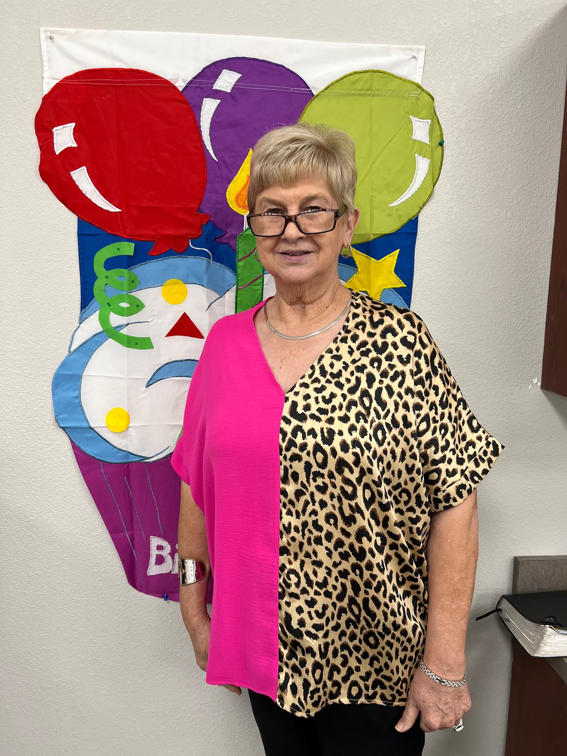 Woman stands in front of a birthday banner. She wears a pink and animal print top, and glasses.