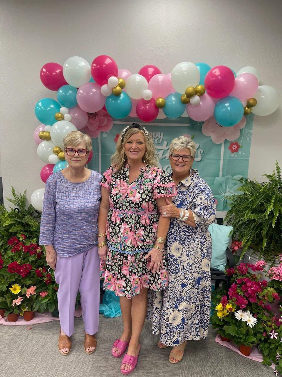 Three women pose in front of a balloon arch and floral display.