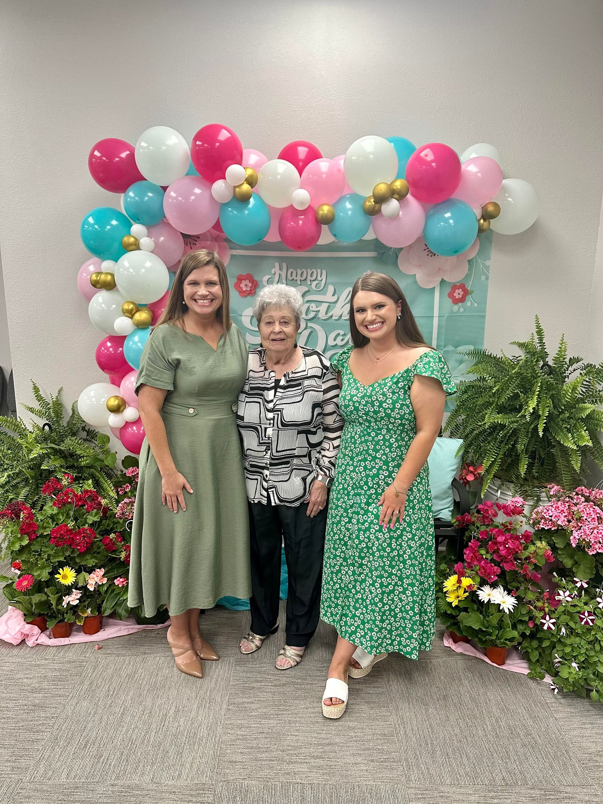 Three women pose in front of a balloon arch; flowers on either side.