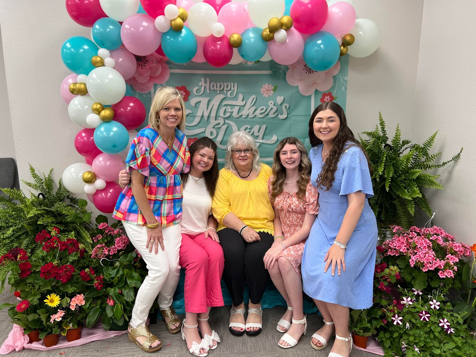 Five women pose for a Mother's Day photo in front of a balloon arch and floral display.