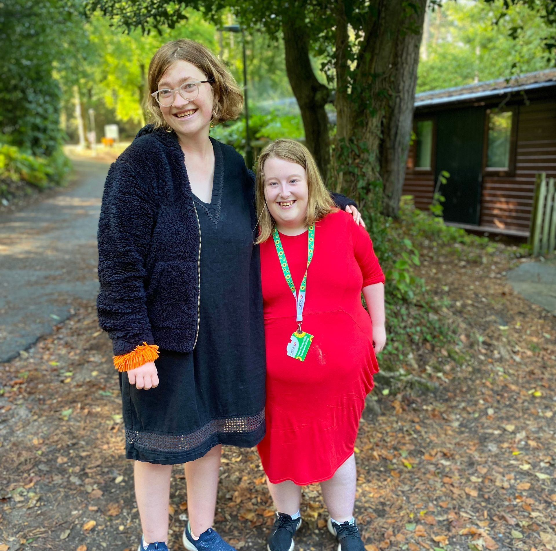 Two young women with Prader-Willi Syndrome hugging standing in front of a wooden lodge and a tree