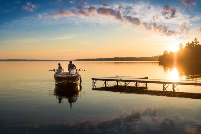 A boat with two people docked on a calm lake at sunrise, with soft golden light reflecting on the water.