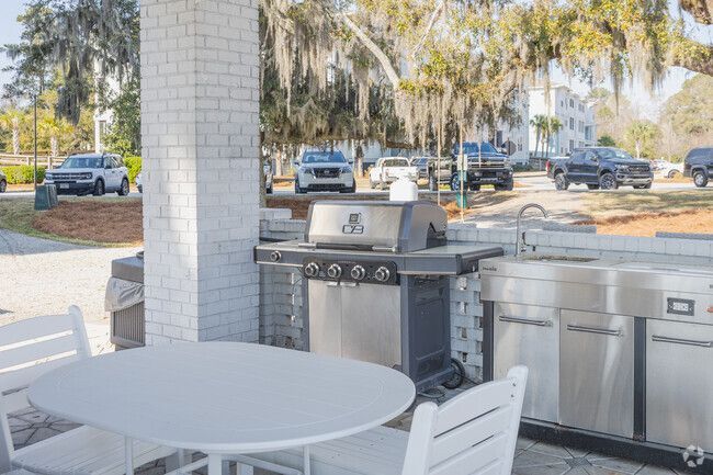 An outdoor grilling station with a metal grill, sink, and white dining table under a covered patio near a parking lot.