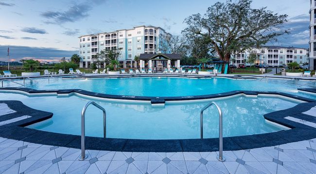 A turquoise swimming pool with two metal handrails in the foreground, set before modern apartment buildings at dusk.