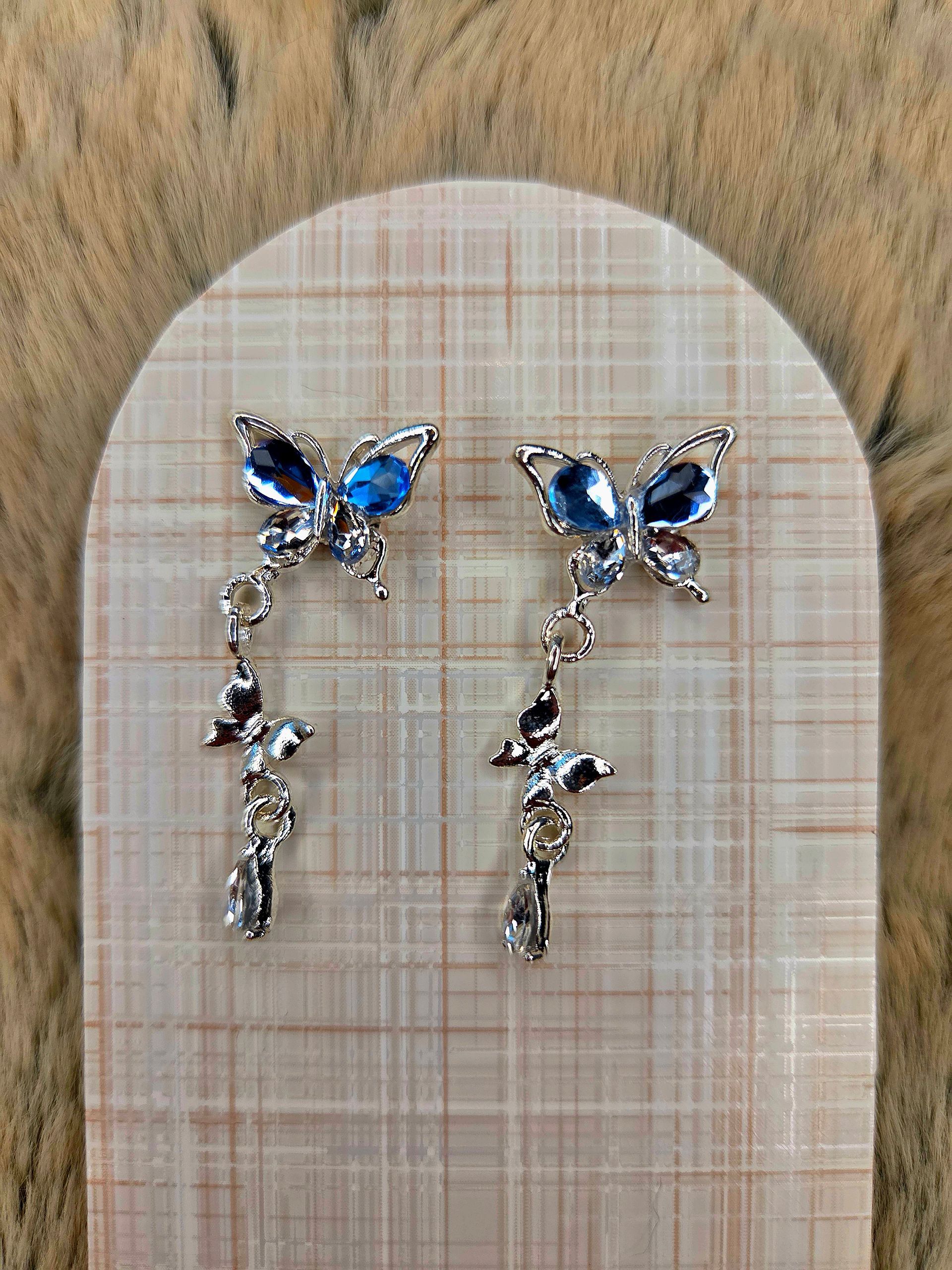 Een paar zilveren oorbellen in de vorm van vlinders met blauwe steentjes op een beige geruite stoffen achtergrond.