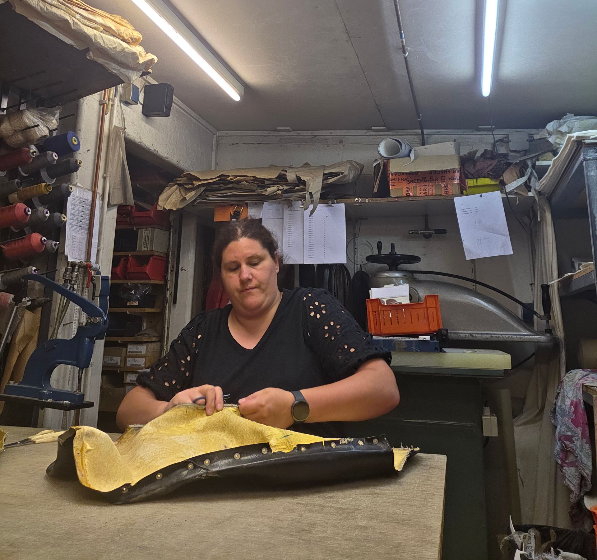 Vrouw in een werkplaats die gele stof knipt op een tafel, met gereedschap en planken op de achtergrond.