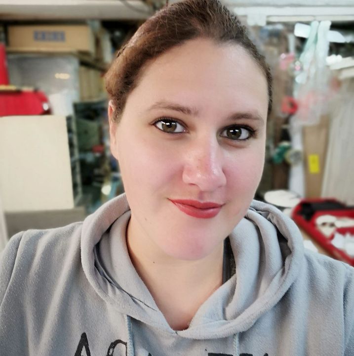 Vrouw met bruin haar en rode lippenstift, gekleed in een grijze hoodie, lachend in een rommelige kamer.