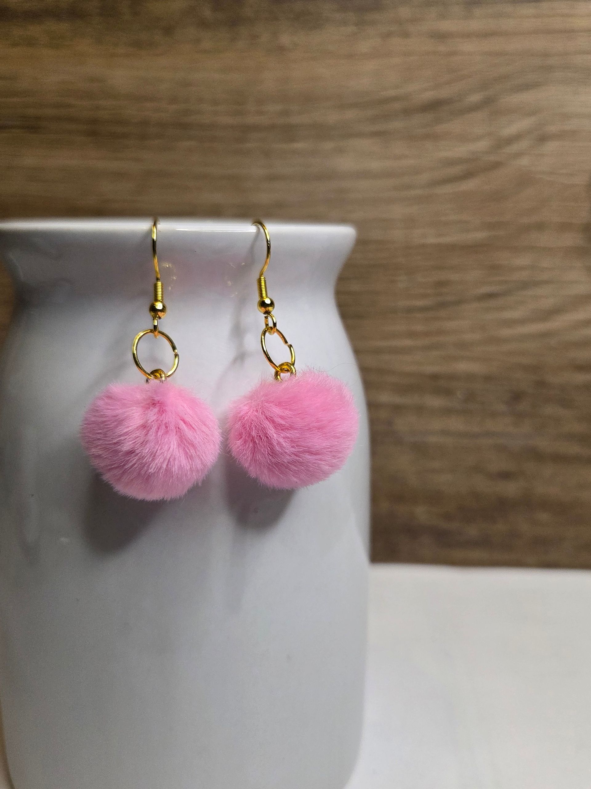 Roze pompon-oorbellen met gouden haakjes hangen aan een witte vaas tegen een houten achtergrond.