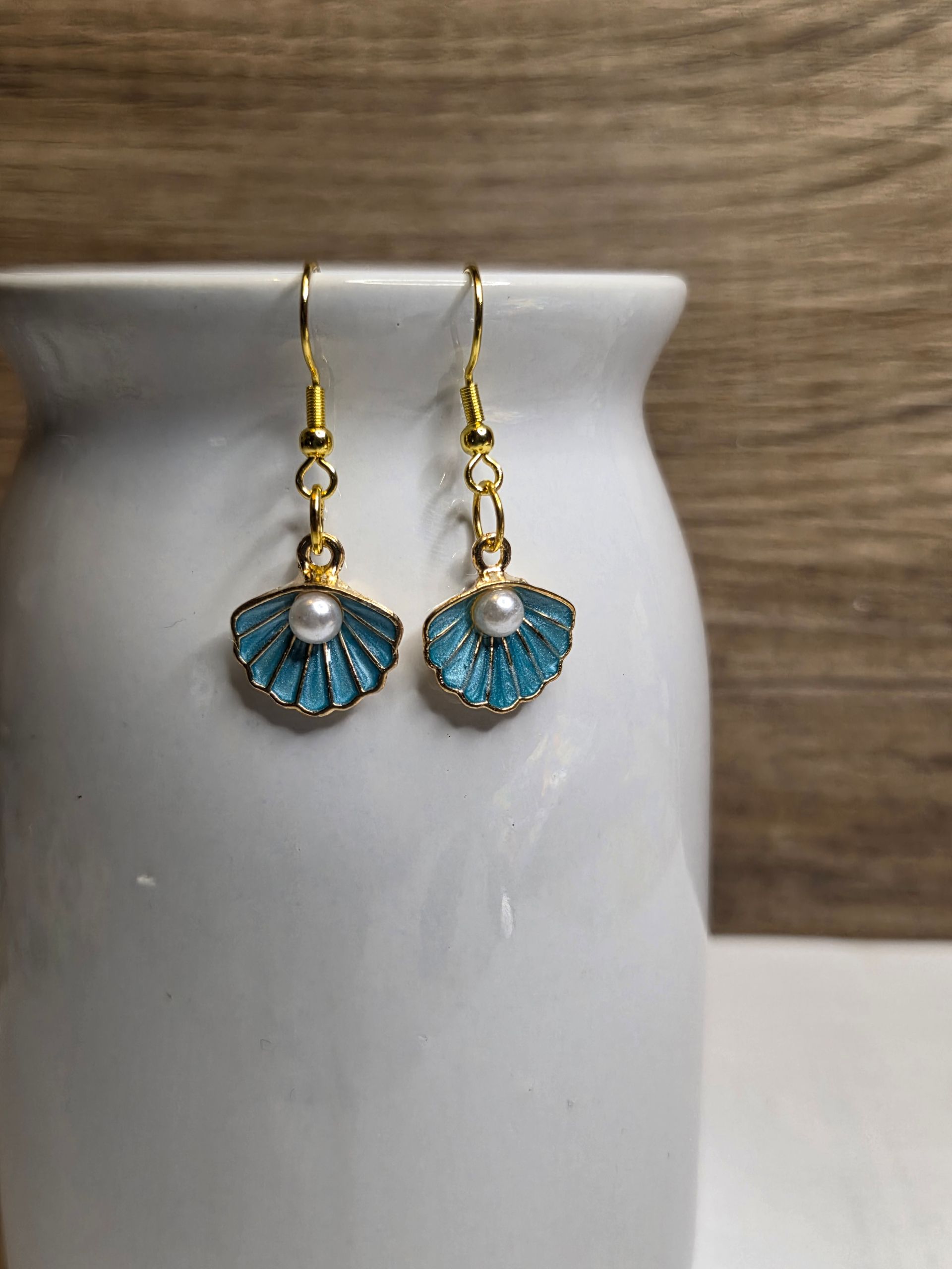 Oorbellen in de vorm van blauwe schelpen met gouden haakjes, gepresenteerd op een witte vaas.