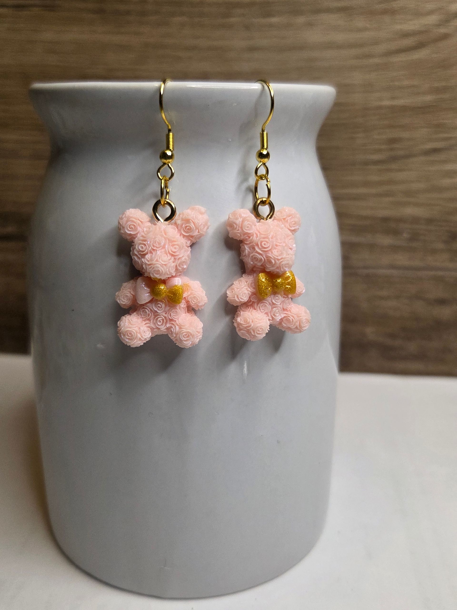 Roze teddybeer-oorbellen met gouden strikjes, hangend aan een grijze vaas tegen een houten achtergrond.