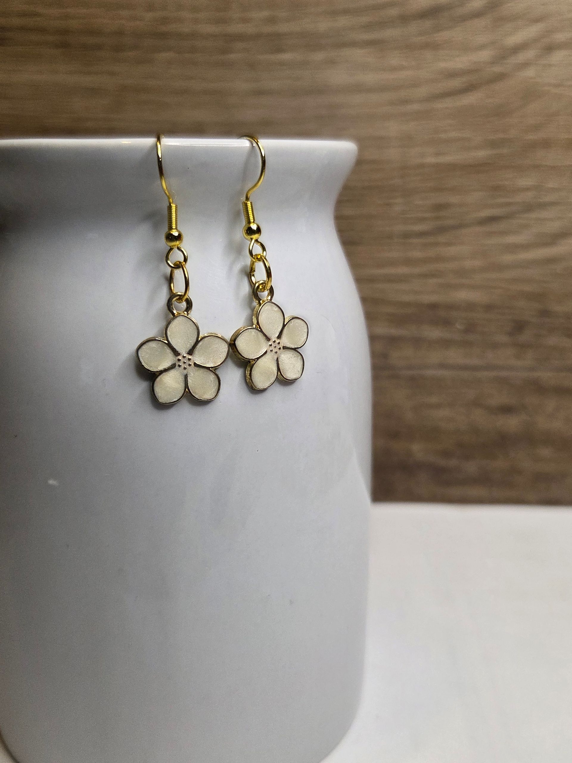 Crèmekleurige bloemoorbellen met gouden haakjes hangen aan een witte keramische pot tegen een wazige houten achtergrond.