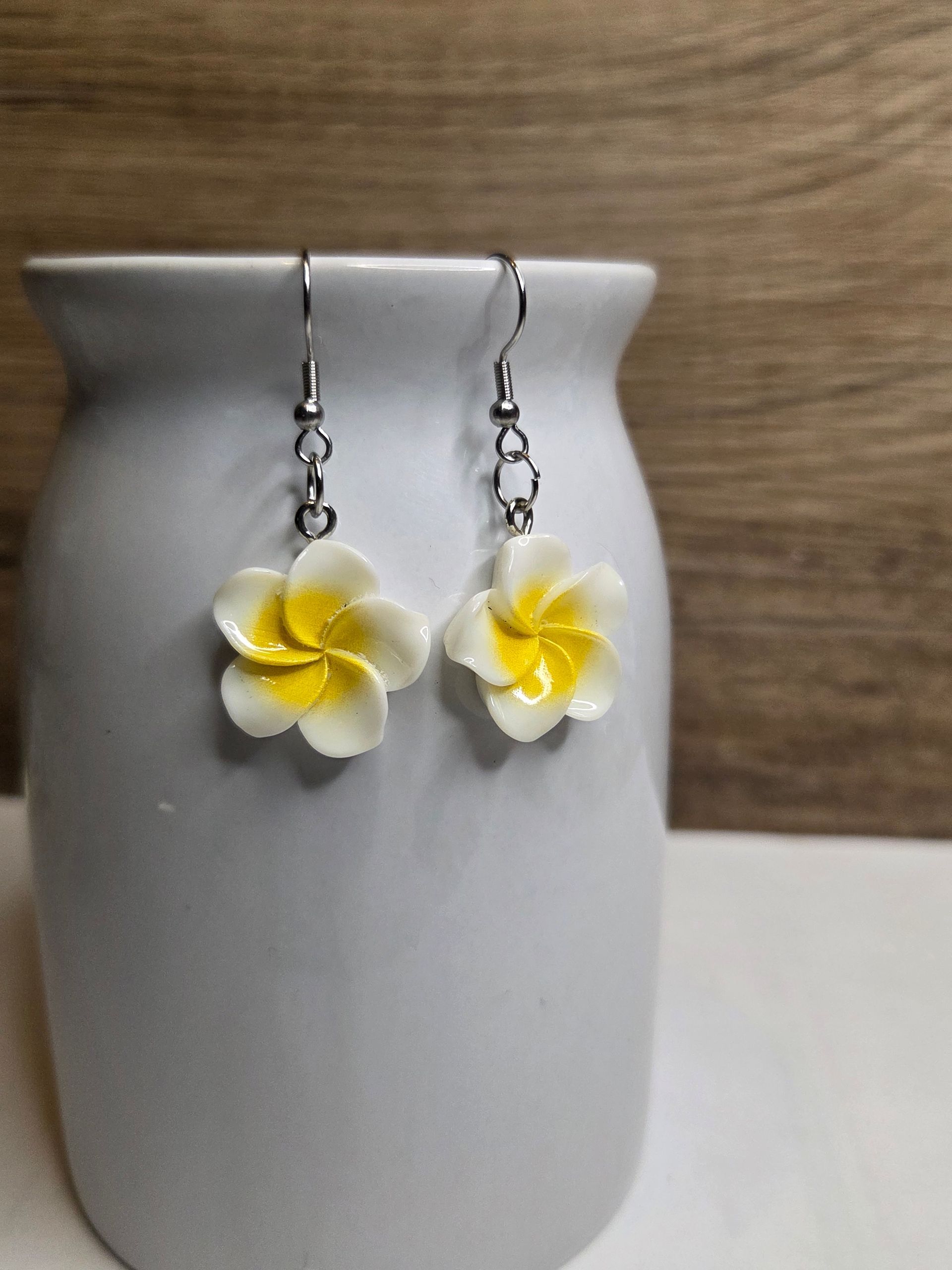 Oorbellen met witte en gele bloemen, hangend aan een witte keramische vaas, tegen een wazige houten achtergrond.