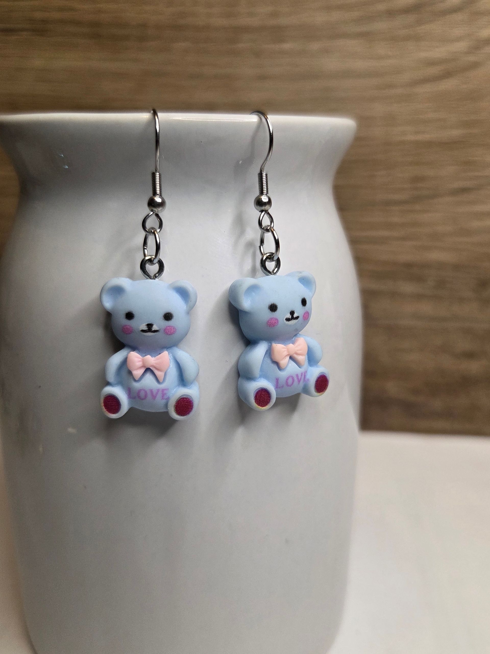 Twee lichtblauwe oorbellen in de vorm van een teddybeer met roze strikjes en het woord "love", hangend aan zilveren haakjes.