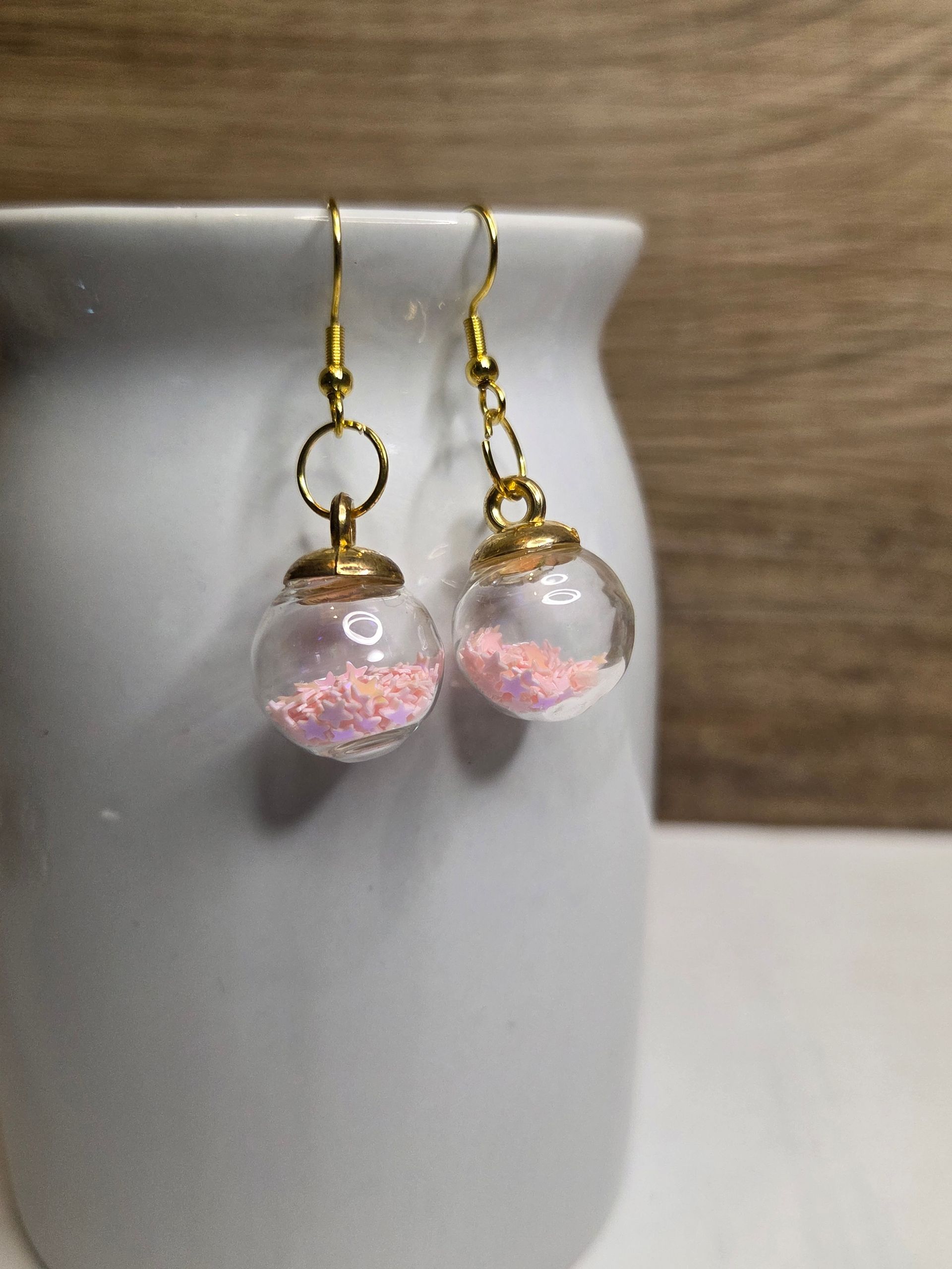 Een paar gouden oorbellen, elk met een transparante bol gevuld met roze glitter, hangend aan een witte houder.