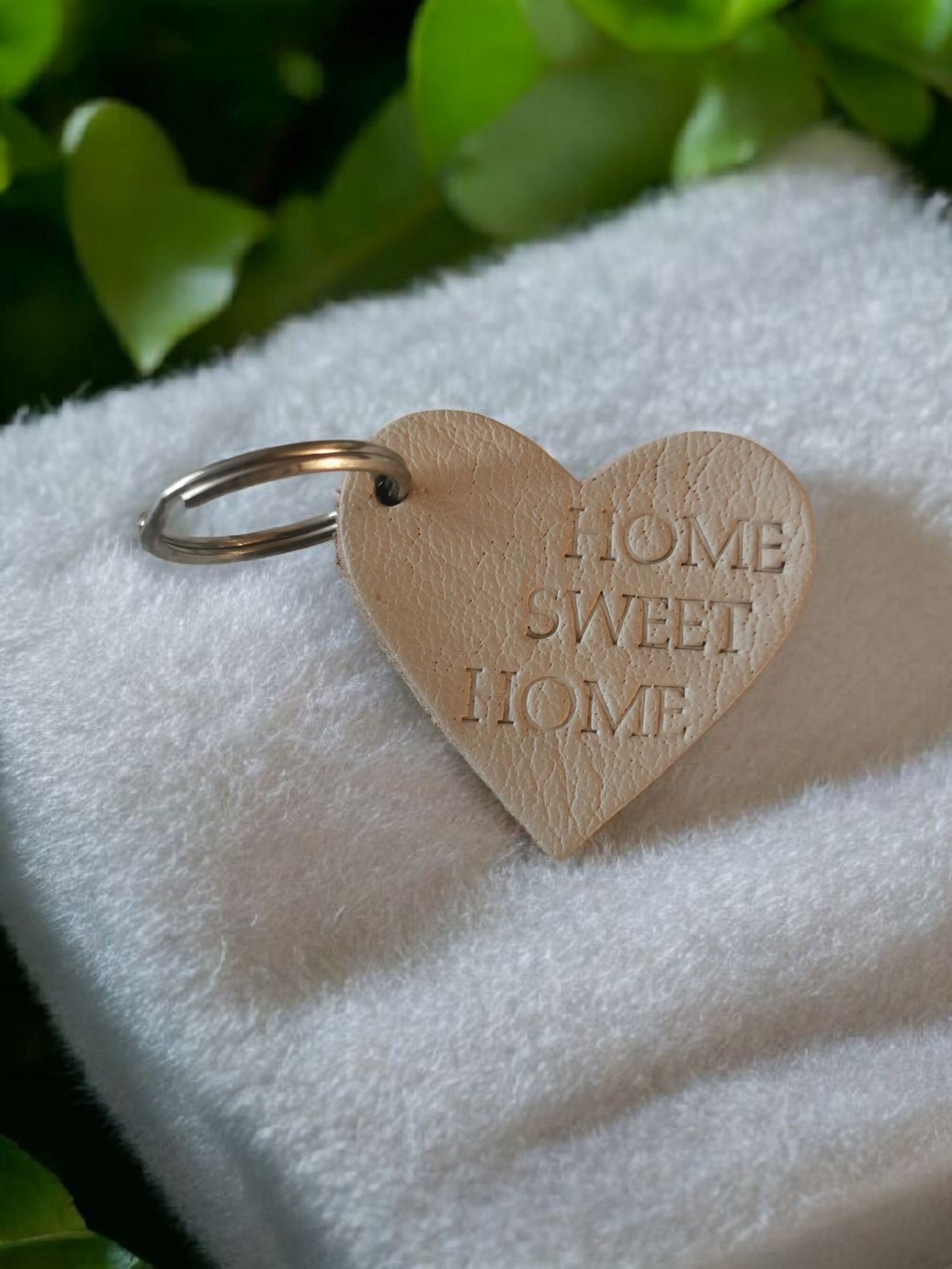 Hartvormige sleutelhanger met de tekst "HOME SWEET HOME" op een witte handdoek met een achtergrond van groene bladeren.