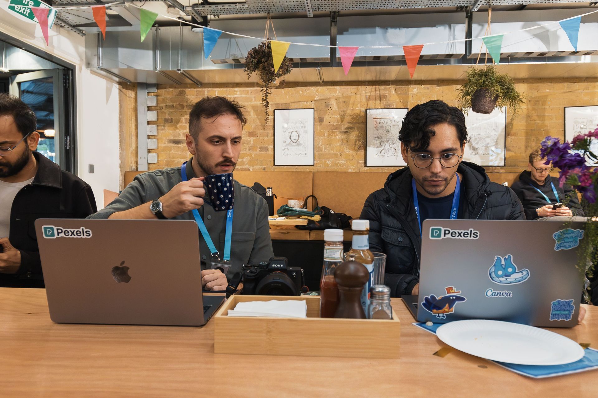 Two people working on laptops at a shared wooden table in a casual office space with bunting overhead