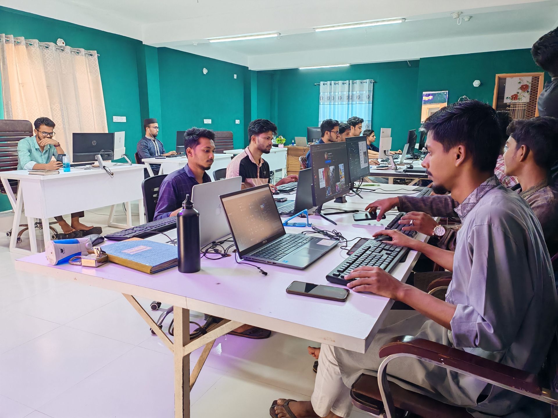 People working at computers in a teal classroom or computer lab with white tables.