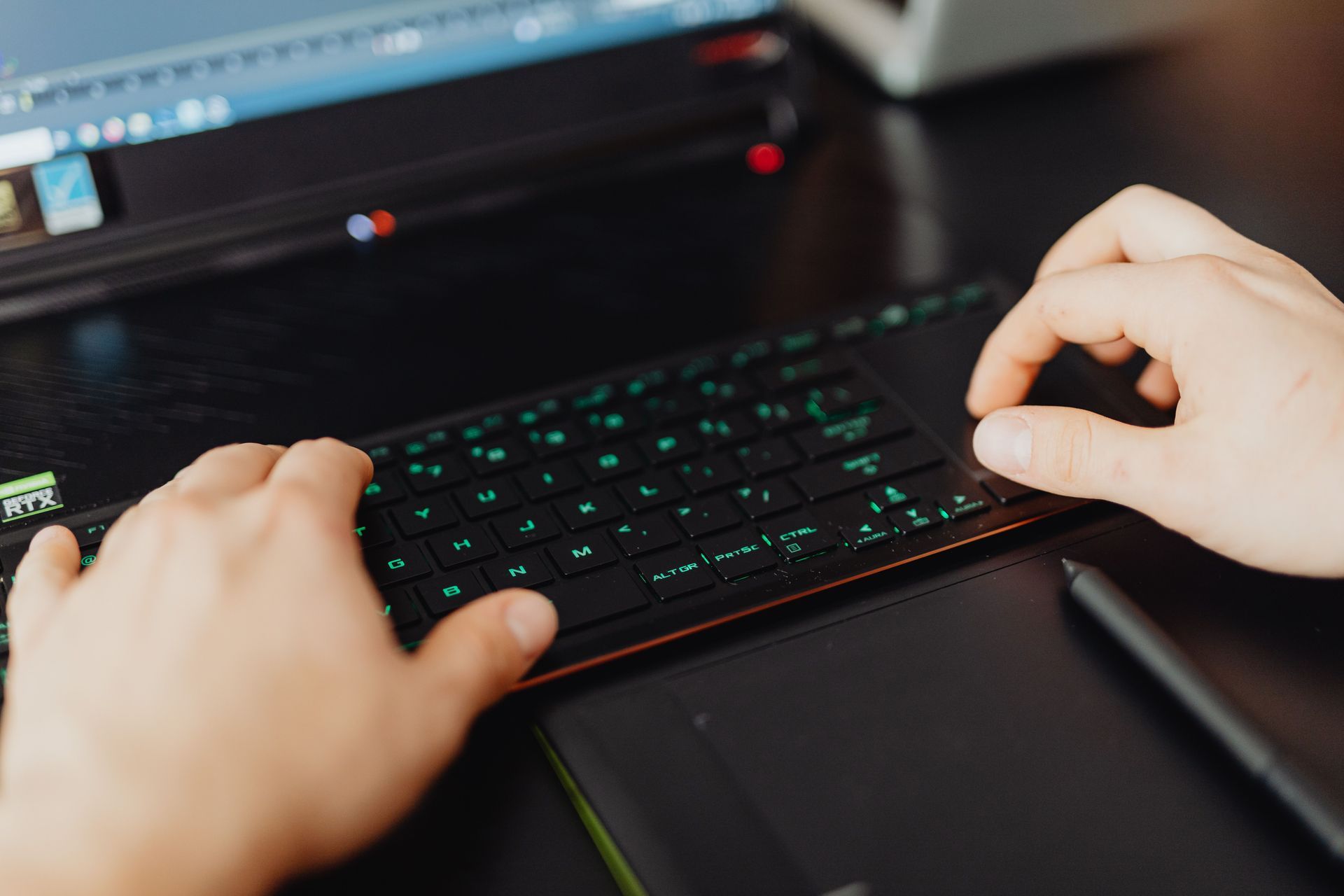 Hands typing on a backlit keyboard beside a laptop and pen on a dark desk