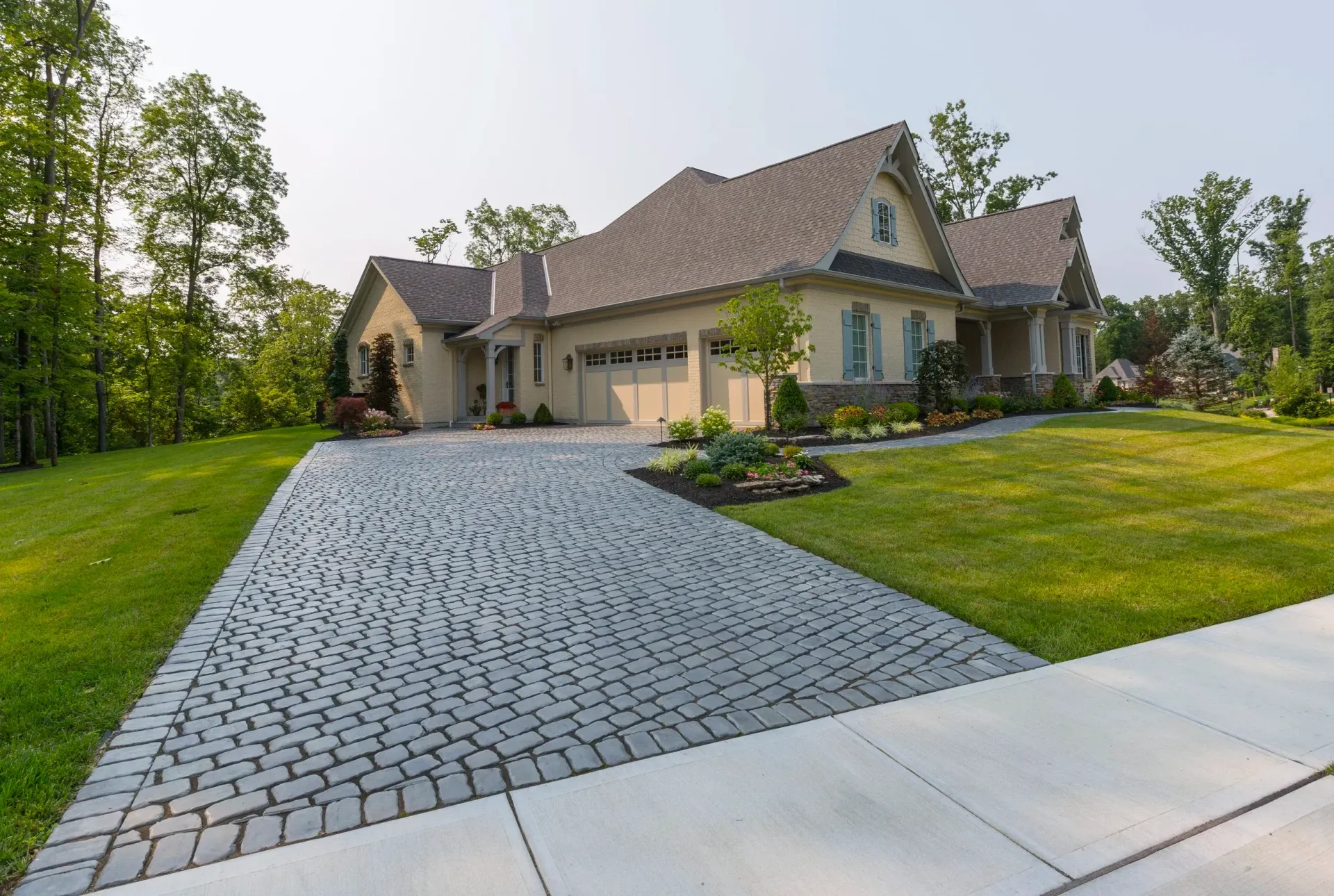 Large suburban house with a cobblestone driveway, manicured lawn, and trees on a sunny day