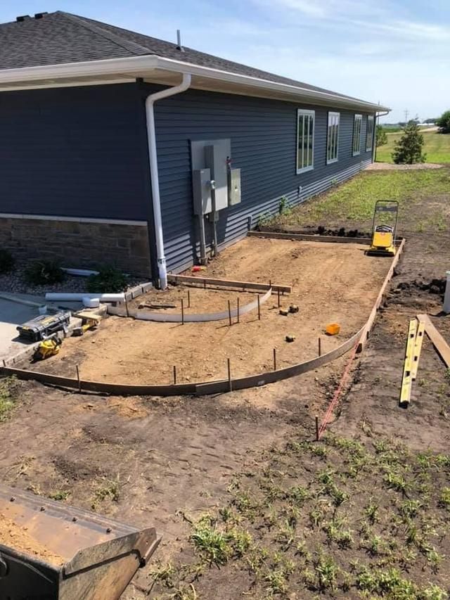Construction site: concrete patio formwork set, beside a blue house with windows, and tan ground.
