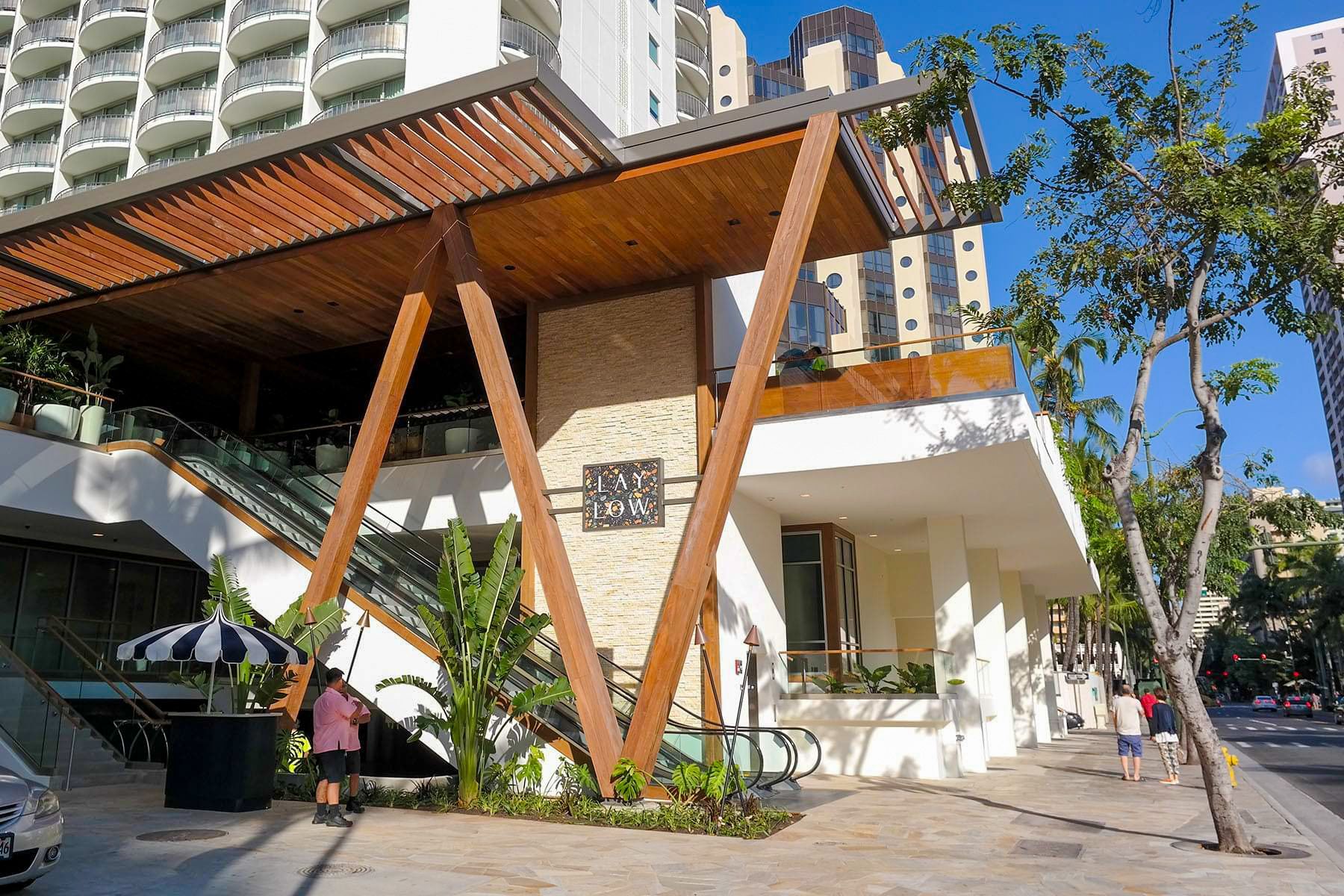 Modern building entrance with wooden canopy, white walls, and trees along a sunny sidewalk