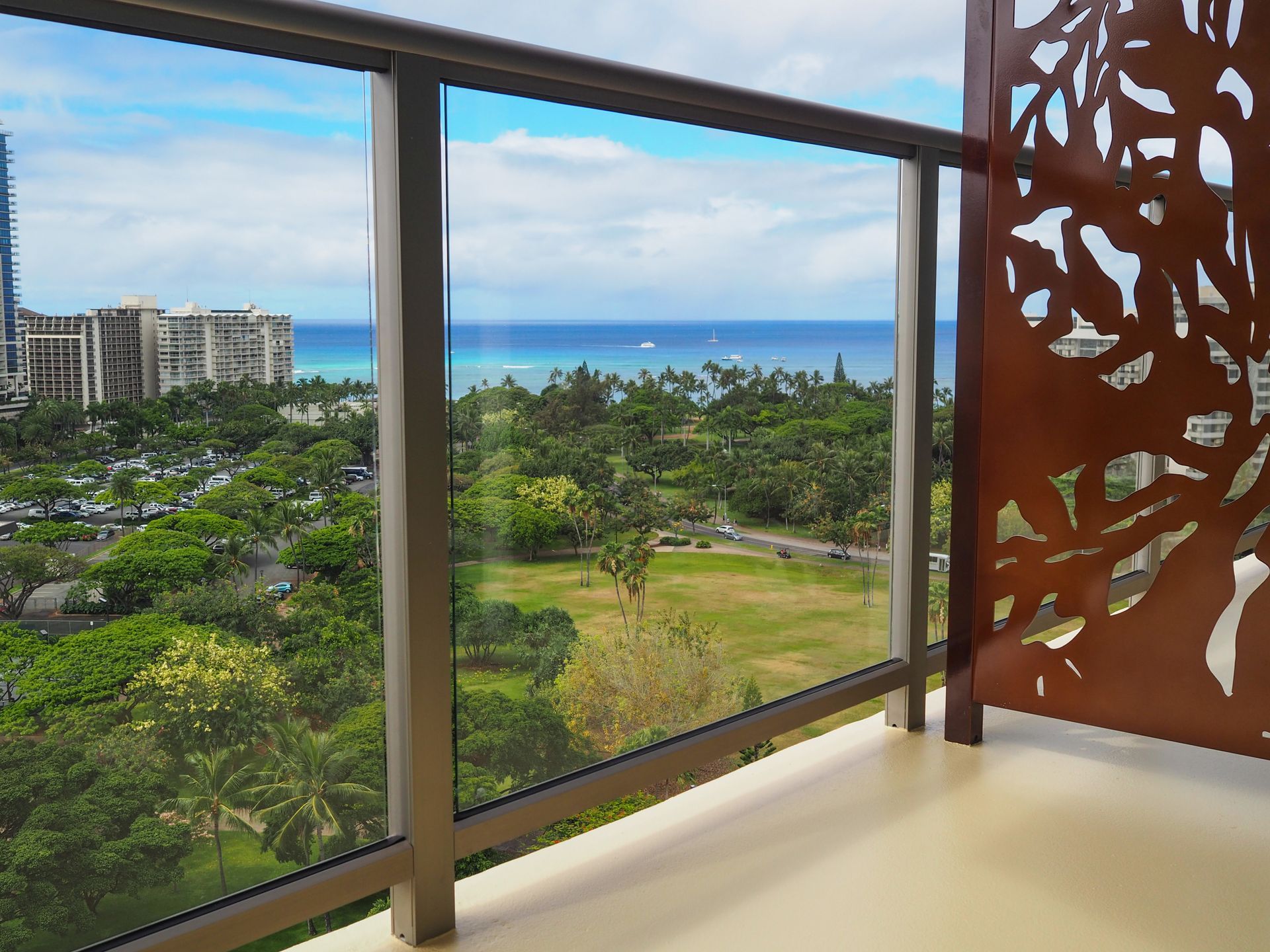 Balcony view of a green park and ocean beyond, framed by glass and a decorative rust-colored panel