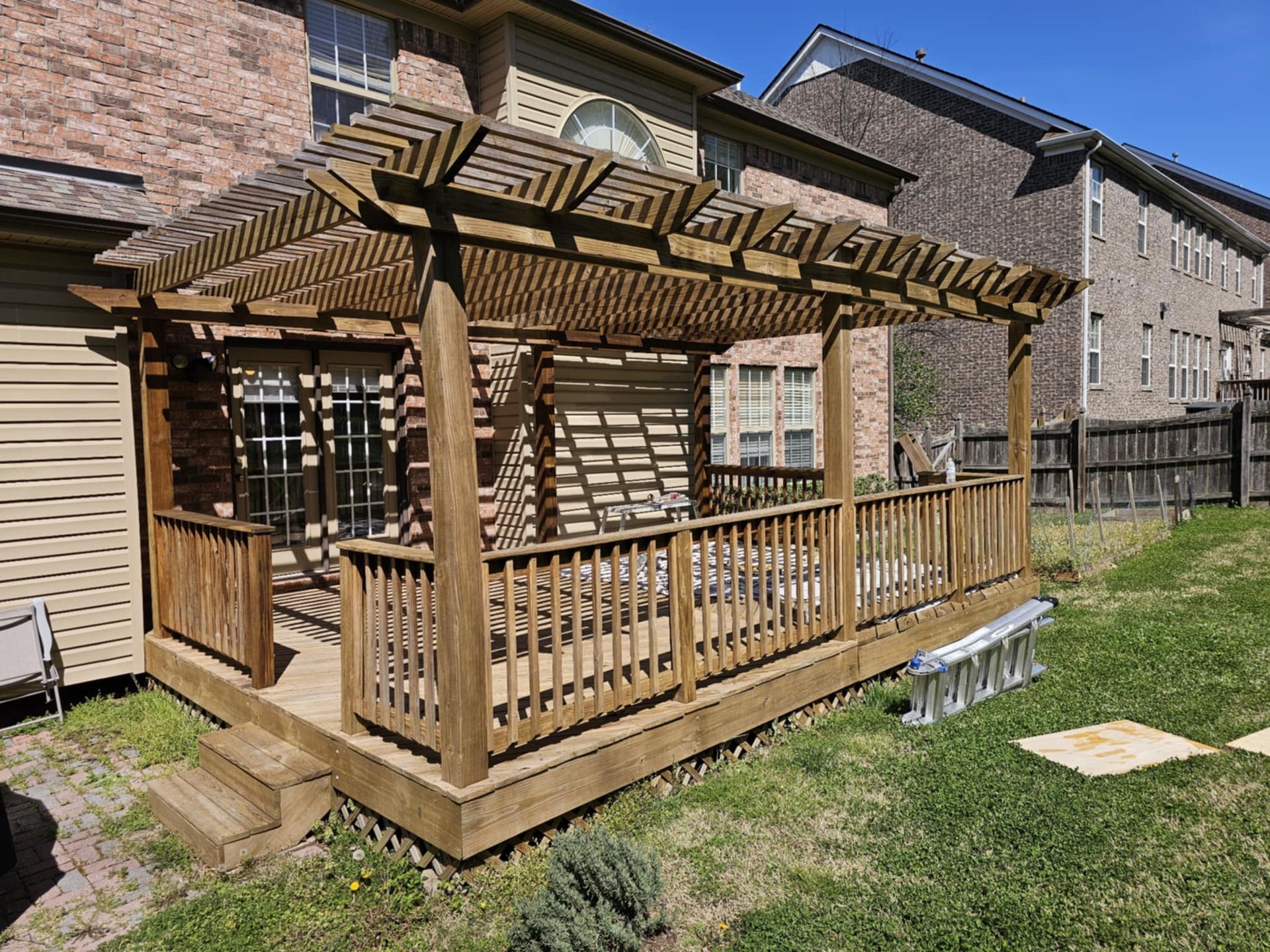 Wooden backyard pergola with railing and lattice roof on a sunlit lawn beside a house