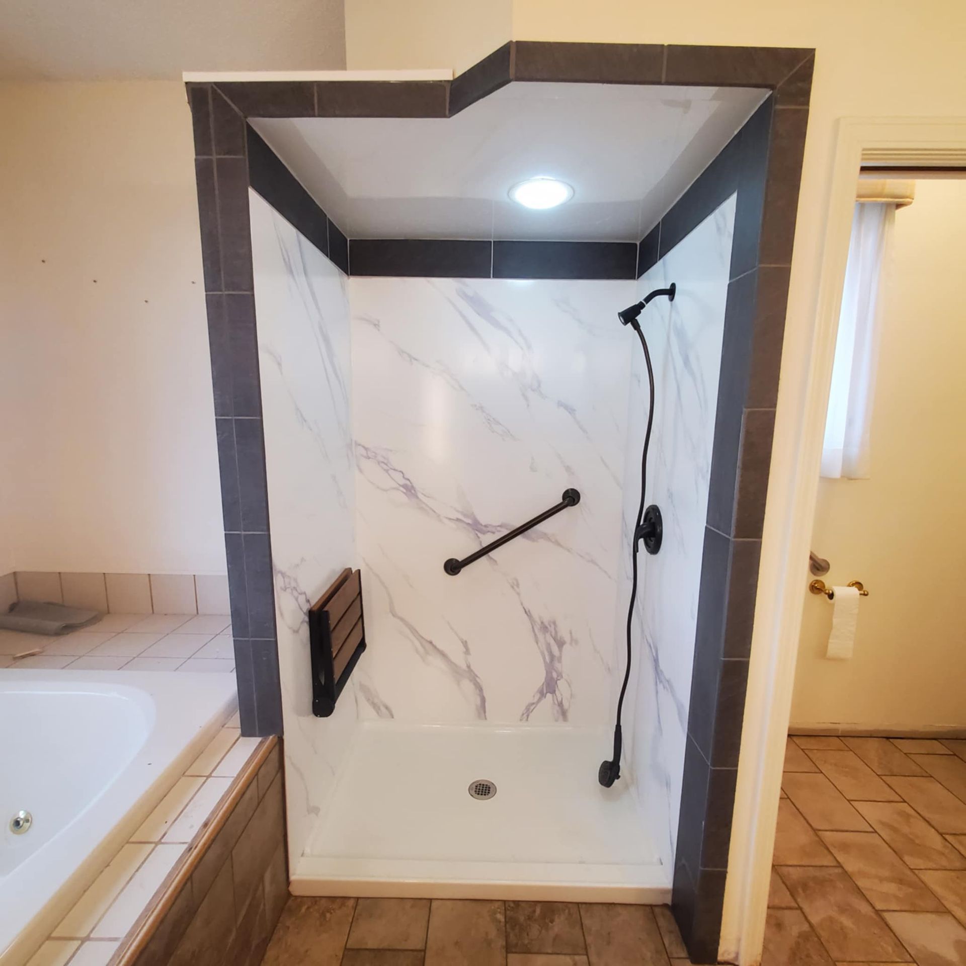 Modern walk-in shower with marble-look tile, dark trim, grab bars, and built-in bench beside a tub