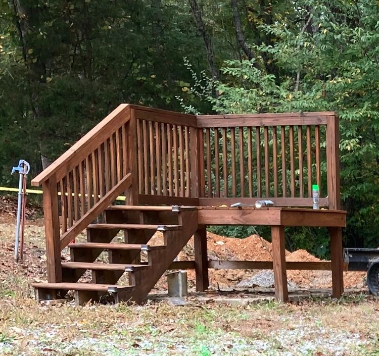 Wooden deck with stairs and railings in a wooded backyard, raised on posts.