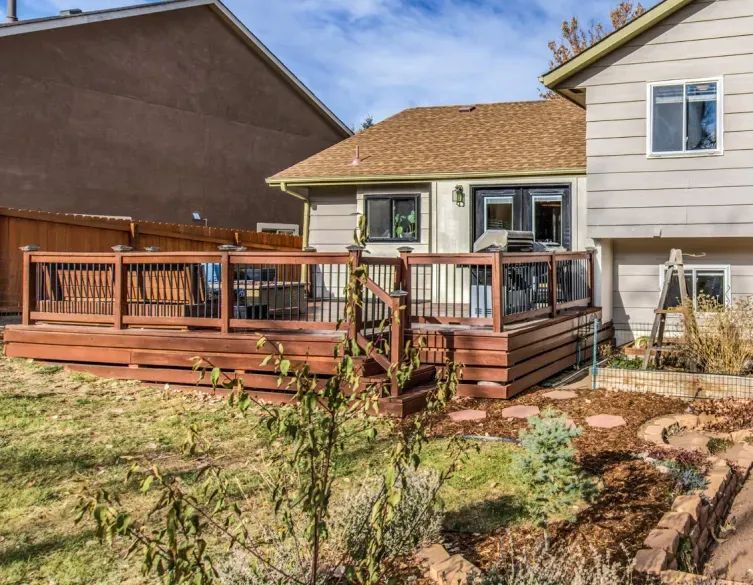 Backyard wooden deck with railing beside a tan house and garden beds