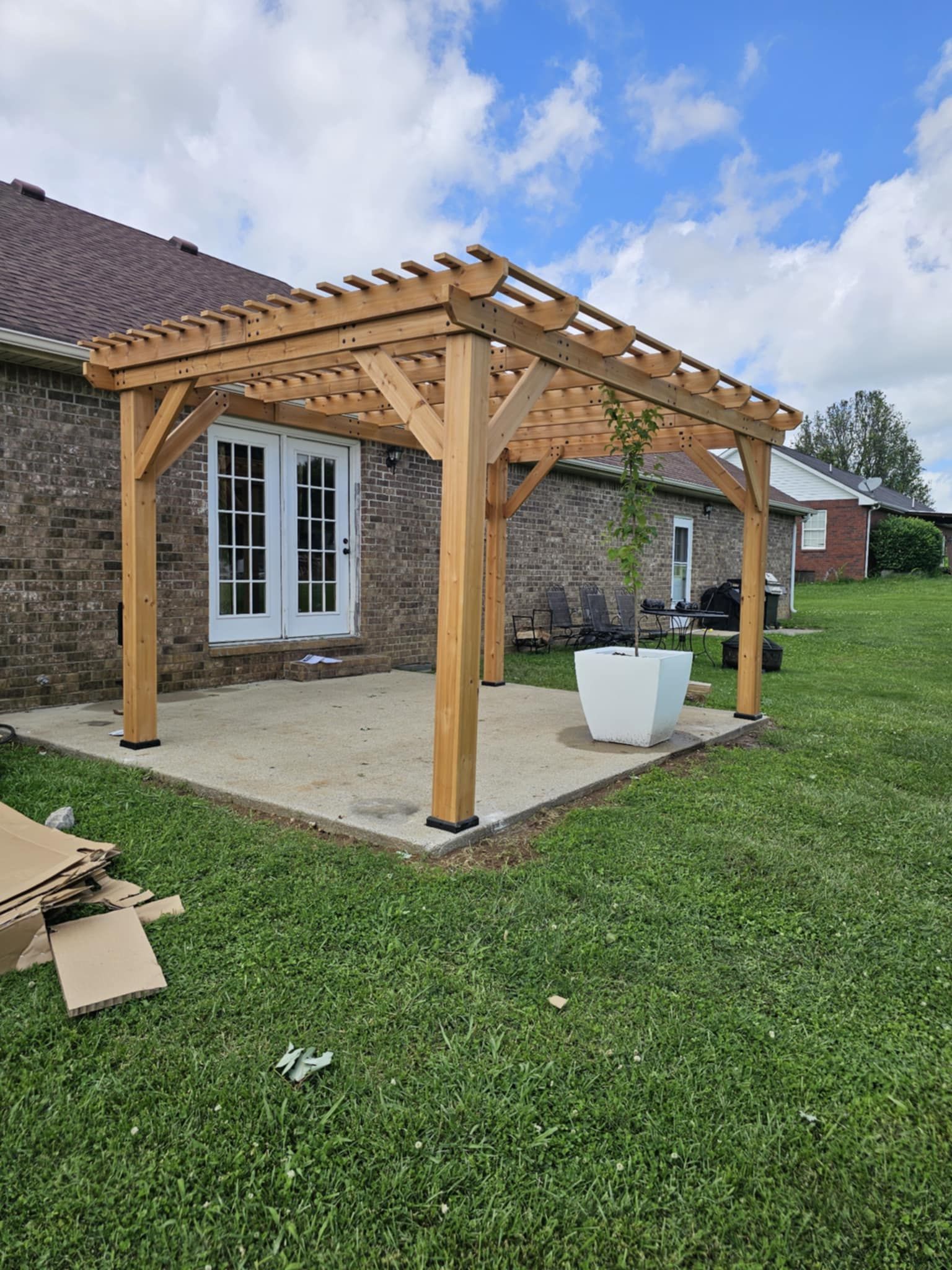 Wooden pergola on a concrete patio beside a brick house, with white planters and a grassy yard.