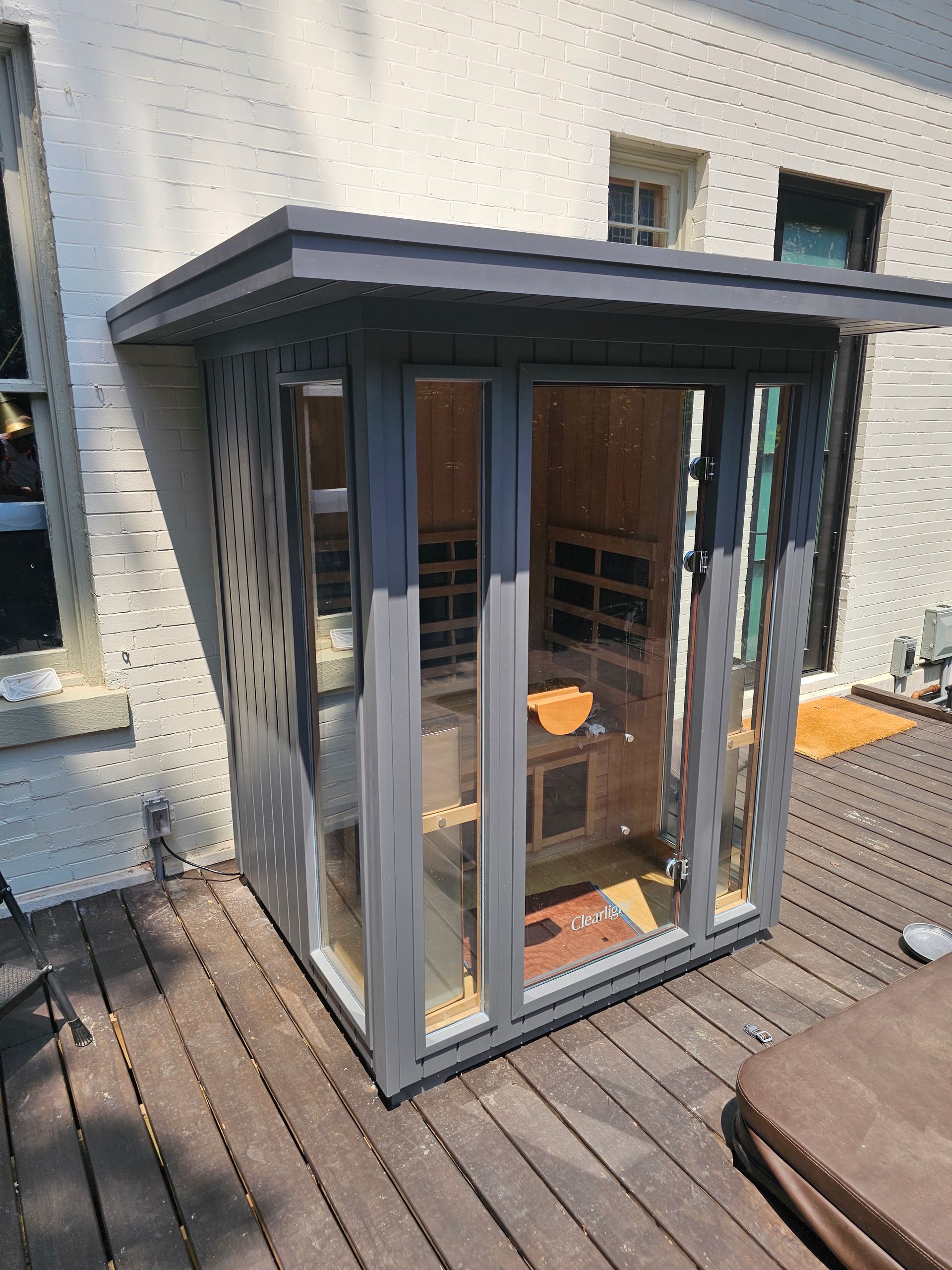 Small glass-front sauna or outdoor cabin on a wooden deck beside a white house