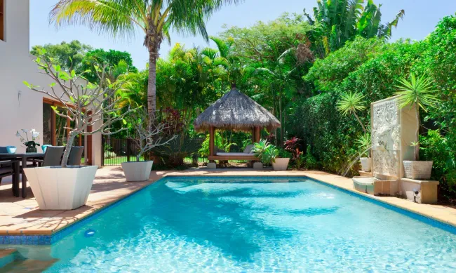 Tropical backyard pool with turquoise water, lounge chairs, palm trees, and a thatched gazebo.