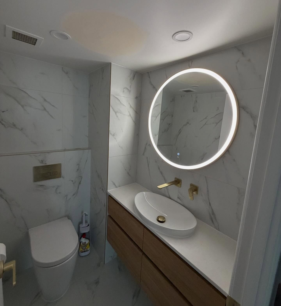 Modern bathroom with marble walls, wall-mounted toilet, floating vanity, and round illuminated mirror