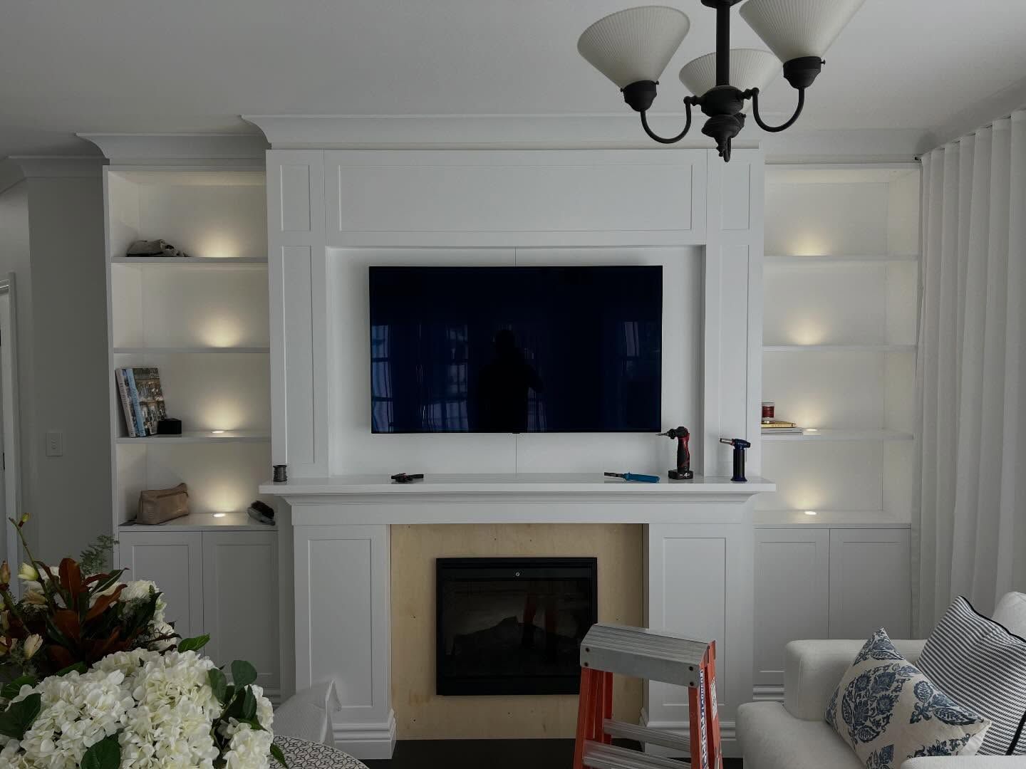 Bright white living room with a fireplace, wall-mounted TV, built-in shelves, and a chandelier