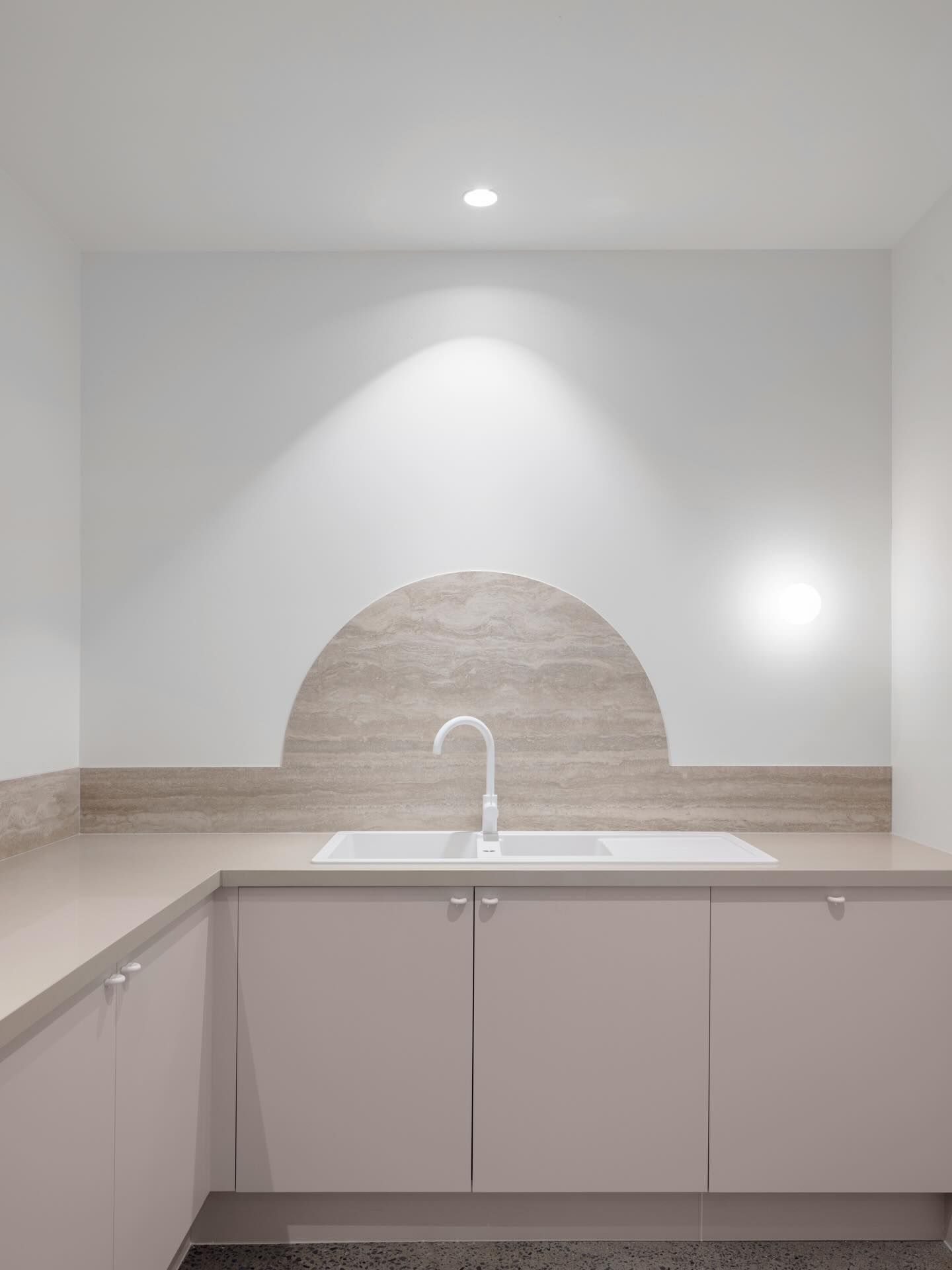 Minimalist kitchen with white walls, beige countertops, and a sink beneath a lit arched backsplash.