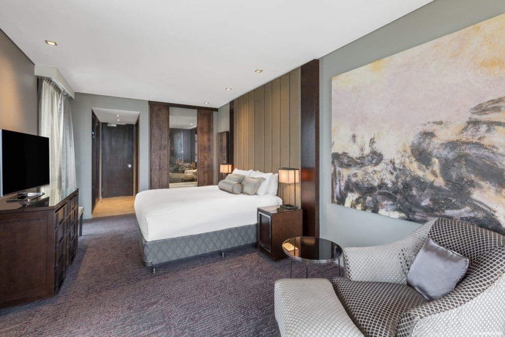 Modern hotel room with bed, armchair, TV, and large wall art in neutral tones