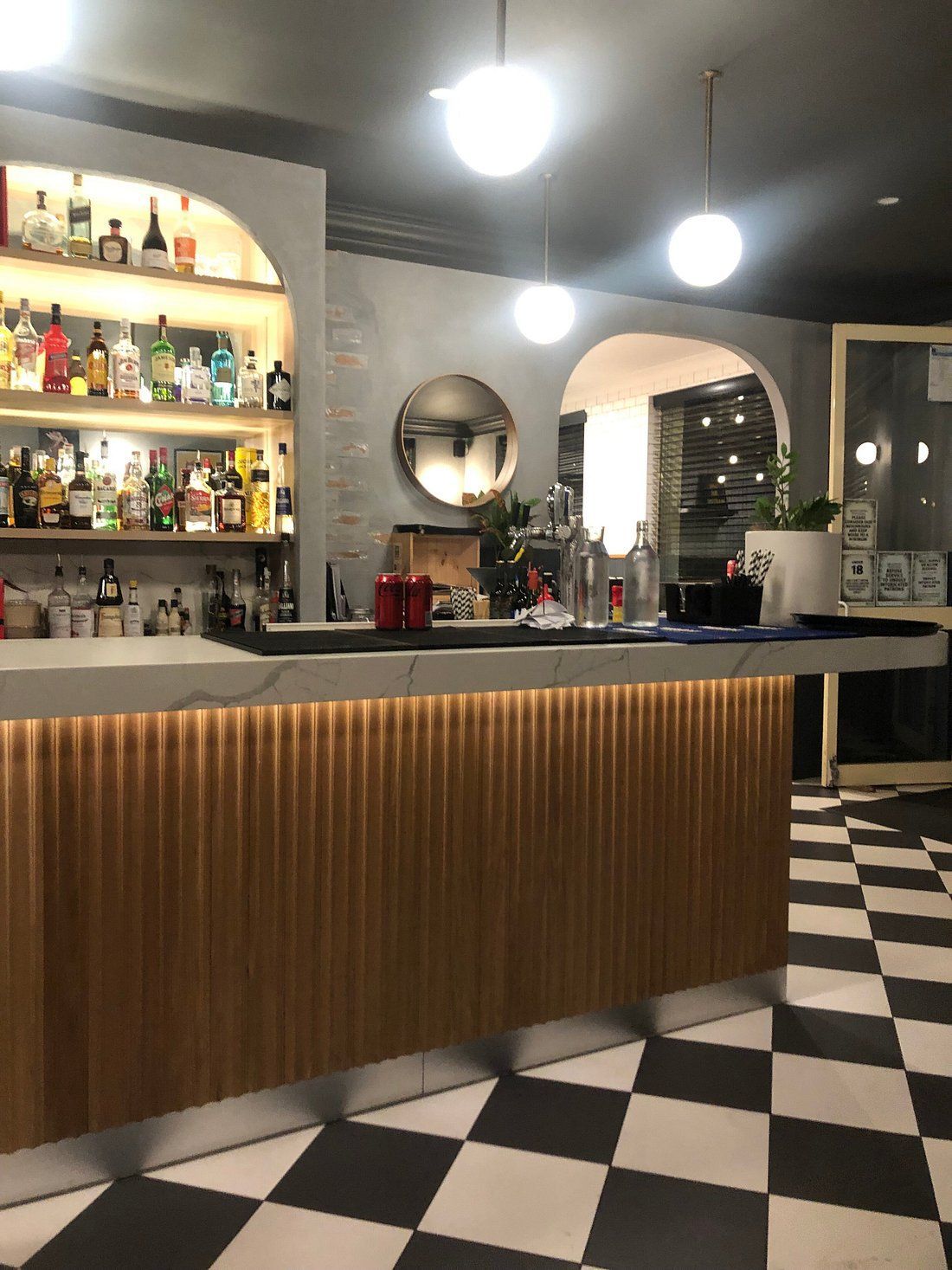 Modern bar interior with curved wooden counter, backlit shelves, and black-and-white checkered floor