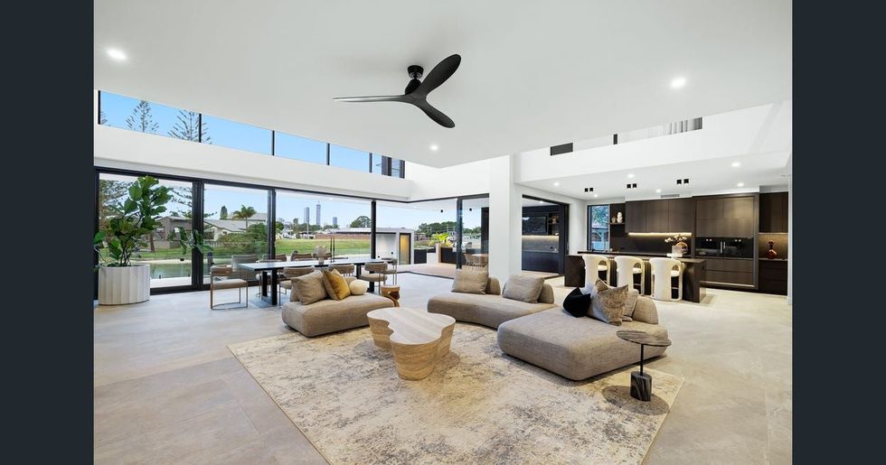 Modern open-plan living room with large windows, beige sectional sofas, and a kitchen area in the background