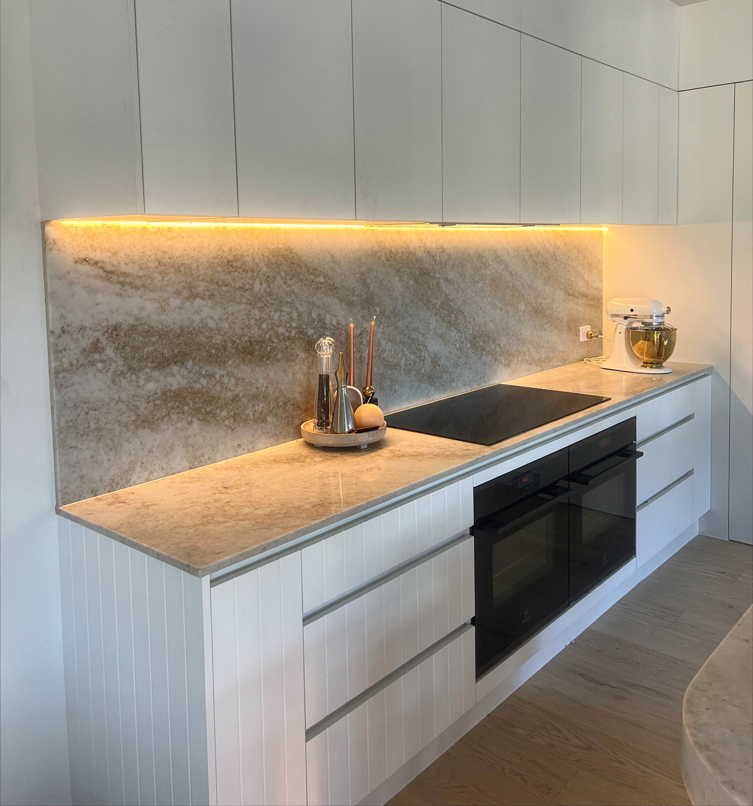 Modern minimalist kitchen with white cabinets, stone backsplash, black oven, and warm under-cabinet lighting