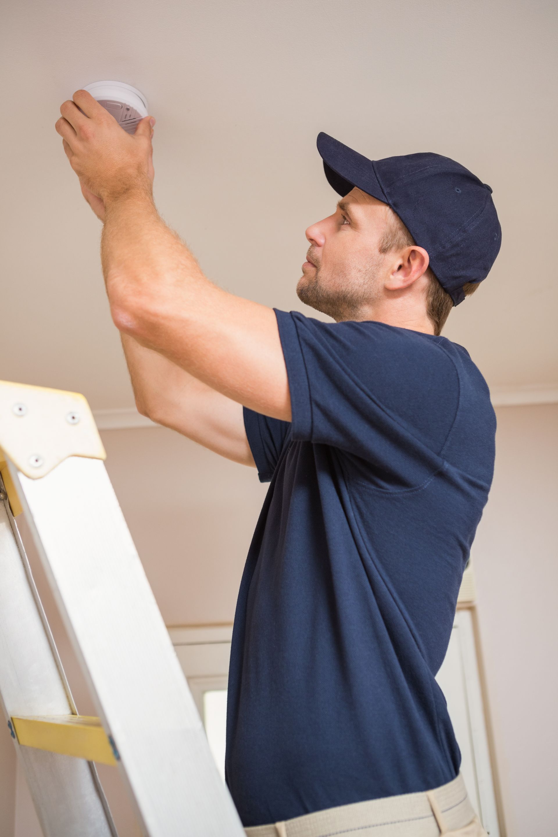 Man in a navy shirt and cap painting a ceiling while standing on a ladder