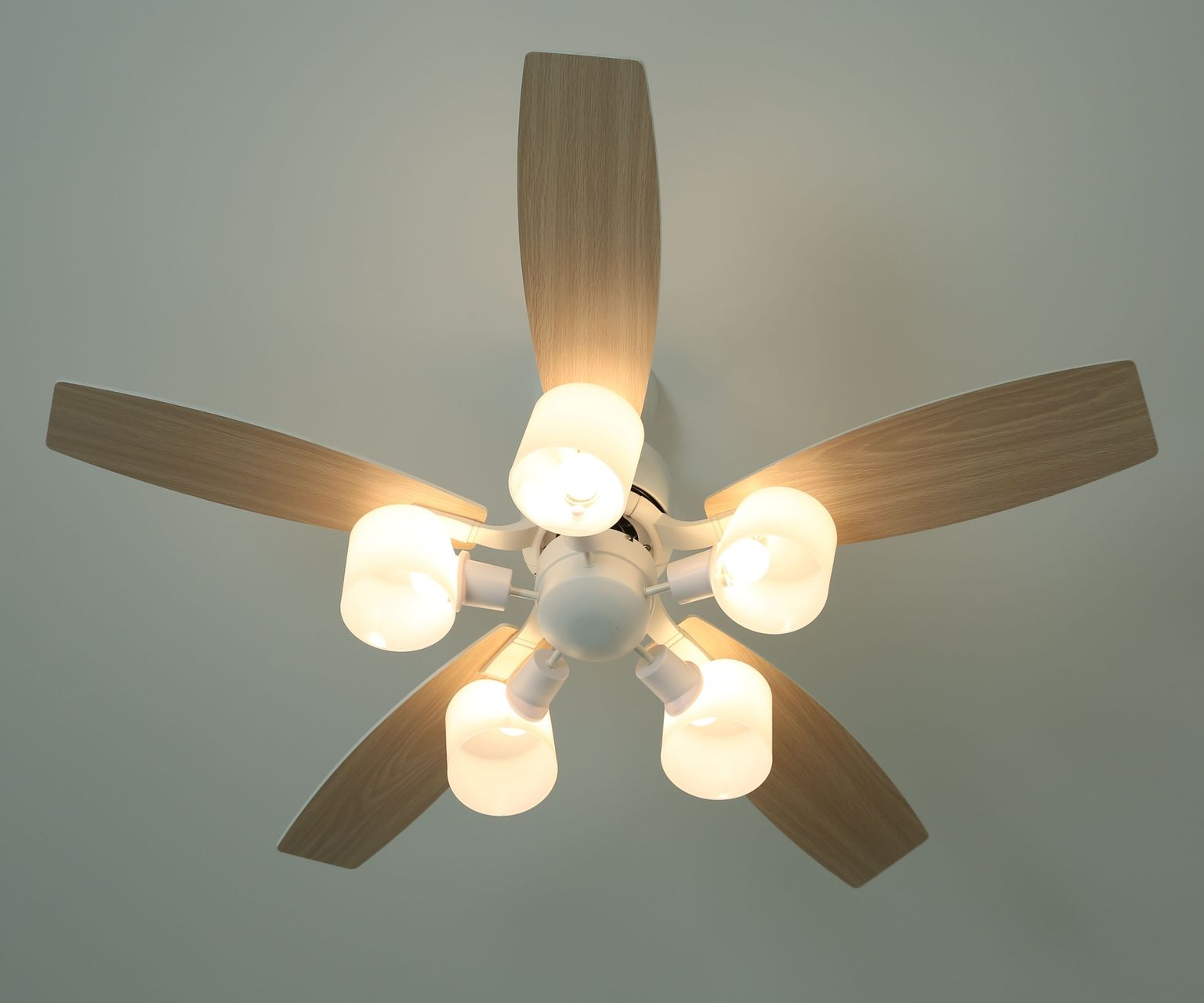 Five-blade ceiling fan with frosted lights and wood-toned blades against a ceiling.