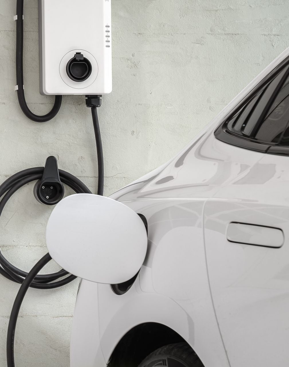 White electric car charging at a wall-mounted charger with cable plugged in.