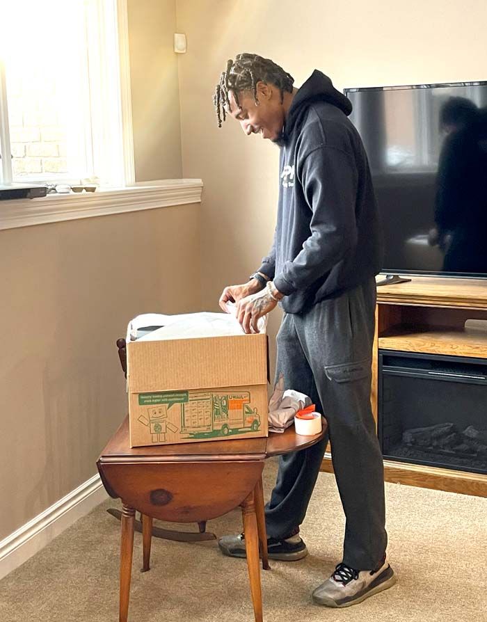 A person in a dark hoodie and sweatpants packs items into a cardboard box on a wooden table in a living room.
