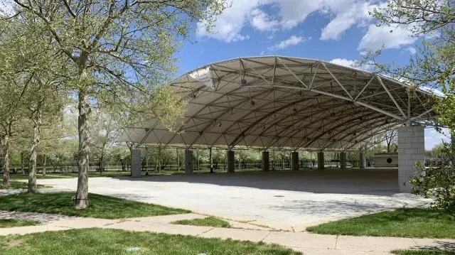 A large, open-air pavilion with a curved metal roof set in a grassy, tree-lined park under a bright blue sky.