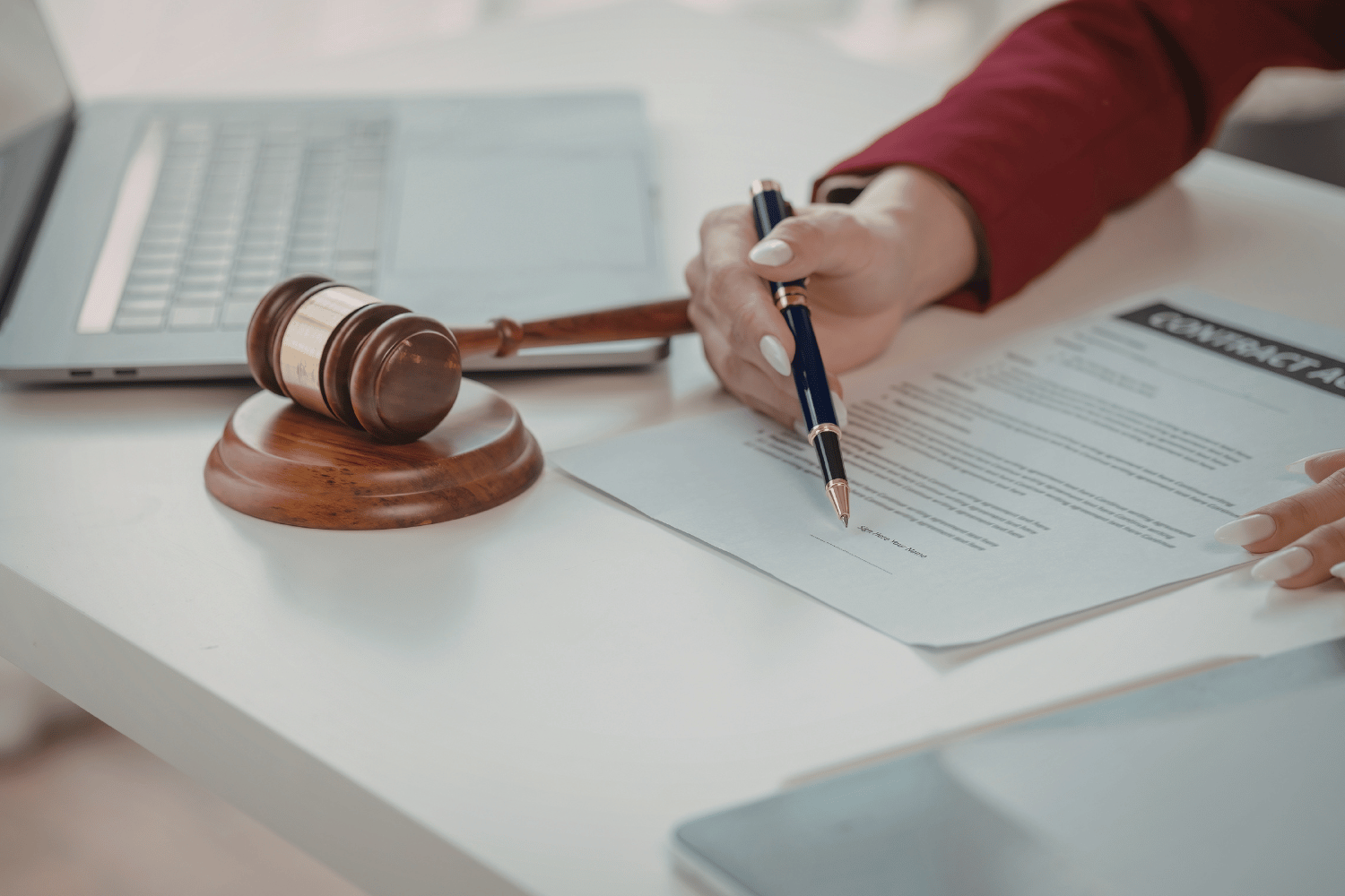 Hand with pen signing a document beside a gavel and laptop on a desk
