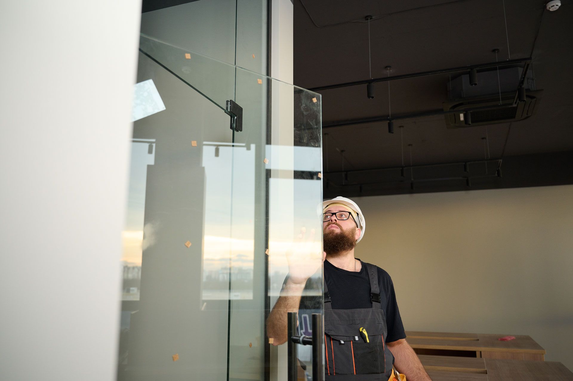 Person in glasses stands by a glass wall inside a modern, unfinished room at sunset.